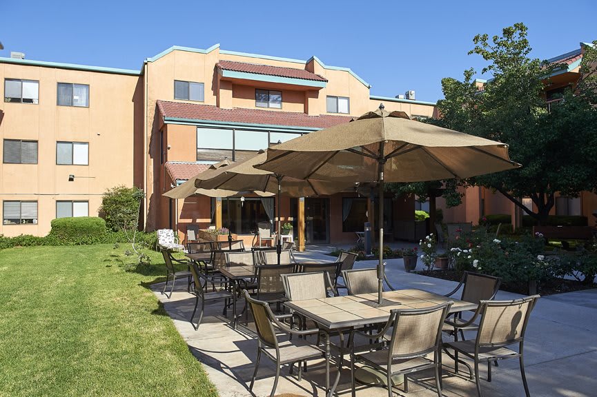 A sunny outdoor patio area with tables, chairs, and umbrellas in front of a building.