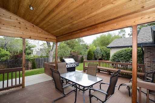 A cozy outdoor patio featuring a dining table, chairs, and a grill, surrounded by lush greenery.