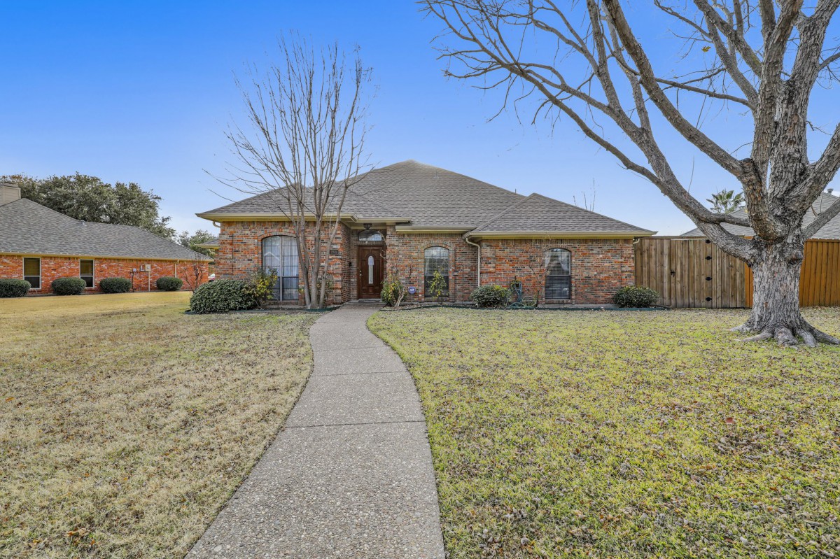 A single-story brick house with a pathway leading to the entrance, surrounded by a lawn and trees.