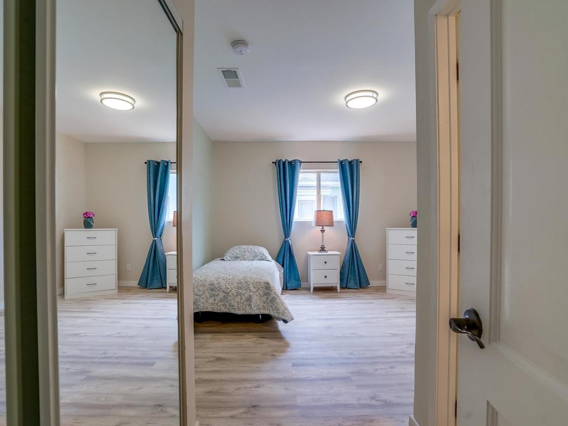 A well-lit bedroom with a single bed, blue curtains, and minimalistic decor reflected in a large mirror.