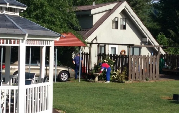 A small house with a garden, a parked car, and people working in the yard.