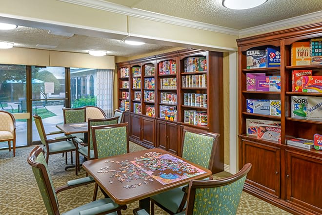 A cozy library with tables and chairs, featuring bookshelves filled with books and puzzles in progress.