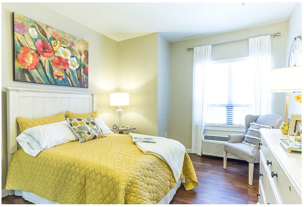 A cozy bedroom with a yellow bedspread, a floral painting above the bed, and a seating area by the window.