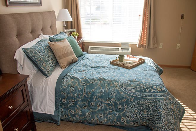 A cozy bedroom featuring a neatly made bed with patterned blue quilt and pillows, a tray on top, and natural light coming through the window.