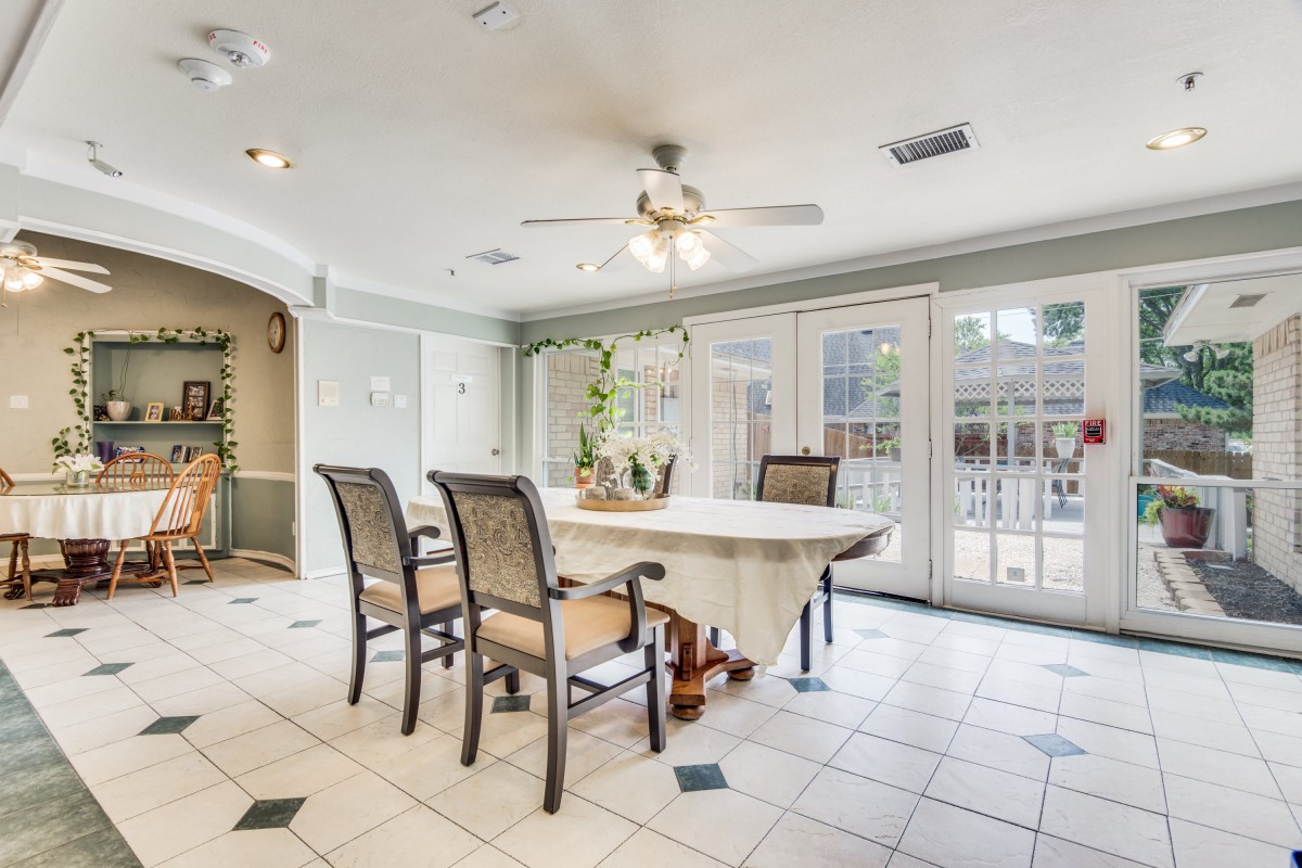 A bright dining room with a large table and wicker chairs, featuring a ceiling fan and ample natural light from the windows.