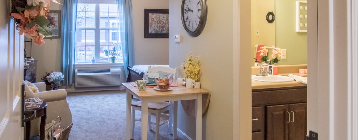 A cozy apartment interior featuring a small table with flowers and decor, a wall clock, and a glimpse of the living room.