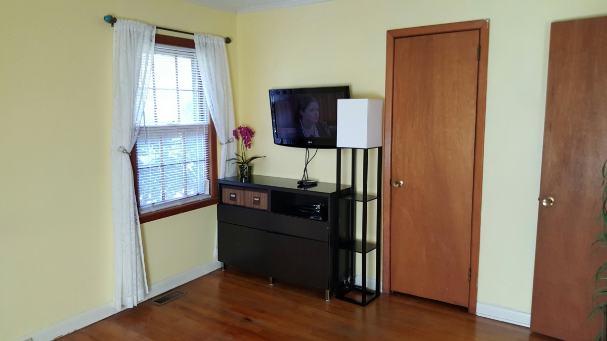 A cozy living room corner with a TV mounted above a dark cabinet, flanked by a modern lamp and adorned with sheer curtains on the window.