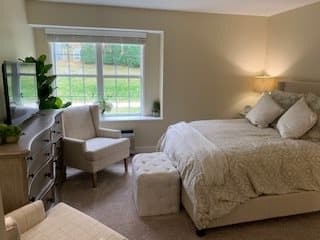 A cozy bedroom with neutral tones featuring a bed, dresser, armchair, and window.