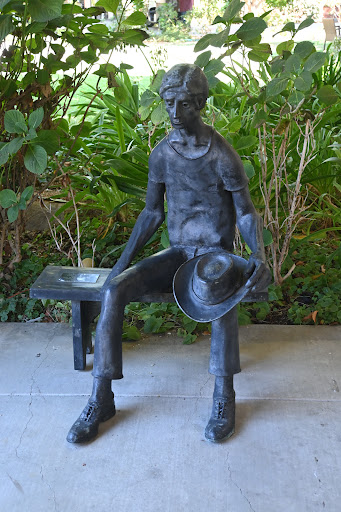 A bronze sculpture of a seated man holding a hat, surrounded by lush greenery.