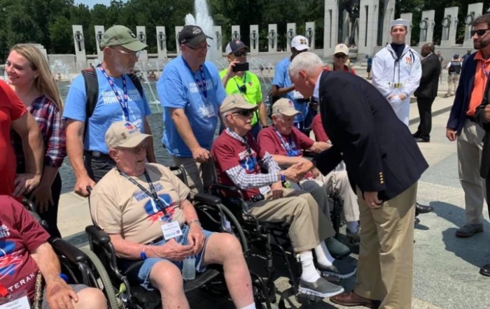 An outdoor gathering where a group of veterans, some in wheelchairs, are being greeted with a handshake.