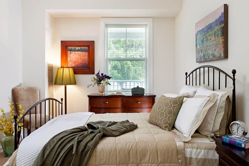 A cozy bedroom featuring a neatly made bed with pillows and a blanket, a window letting in natural light, and a dresser topped with flowers and a lamp.