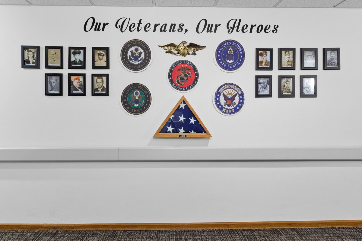 A tribute wall honoring military veterans, adorned with service branch emblems, framed photos, and a folded American flag.