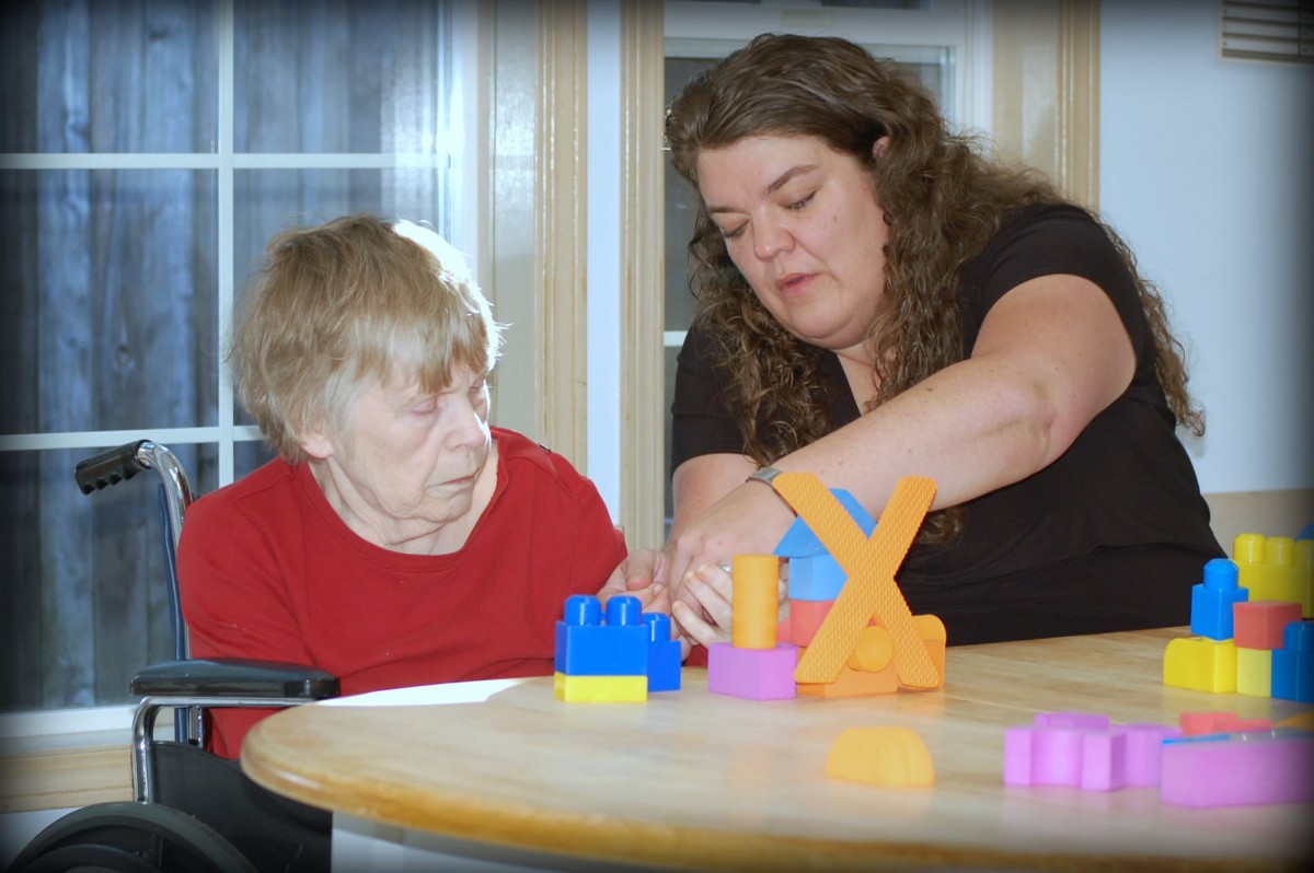 A caregiver assists an elderly person in playing with colorful blocks.