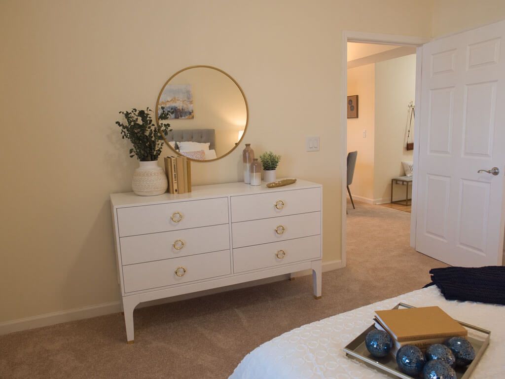 A cozy bedroom featuring a white dresser with a round mirror and decorative plants.