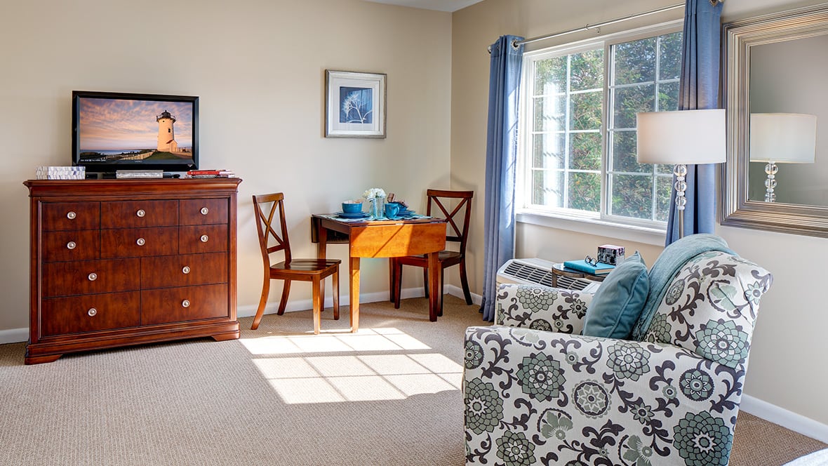 A cozy living room with a table set, chairs, a chest of drawers, and sunlight streaming through the window.