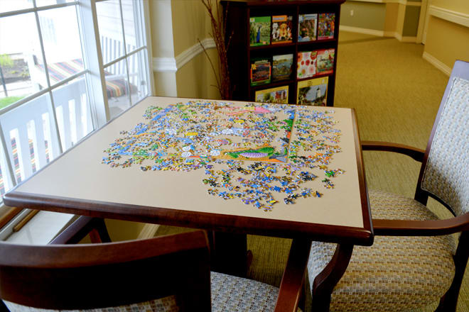 A table with an incomplete jigsaw puzzle near a window, surrounded by chairs and a bookshelf in the background.