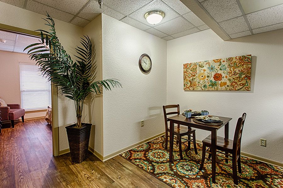 A cozy dining room with a small wooden table set, vibrant rug, decorative wall art, and a potted plant.