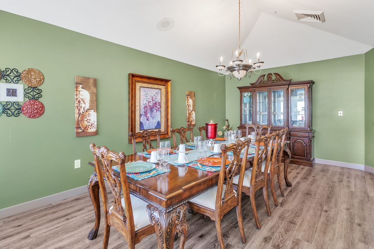 A classic dining room with a large wooden table set for a meal, surrounded by ornate chairs and illuminated by a chandelier. The room features green walls adorned with wall art and decorative plates, as well as a wooden cabinet in the background.