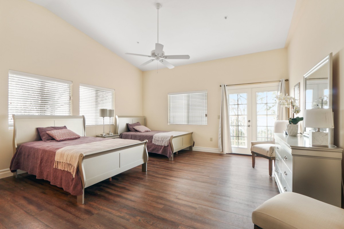 A spacious bedroom featuring two beds, a ceiling fan, hardwood flooring, and large windows.