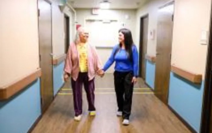 An elderly woman and a young woman walking hand-in-hand down a hallway.