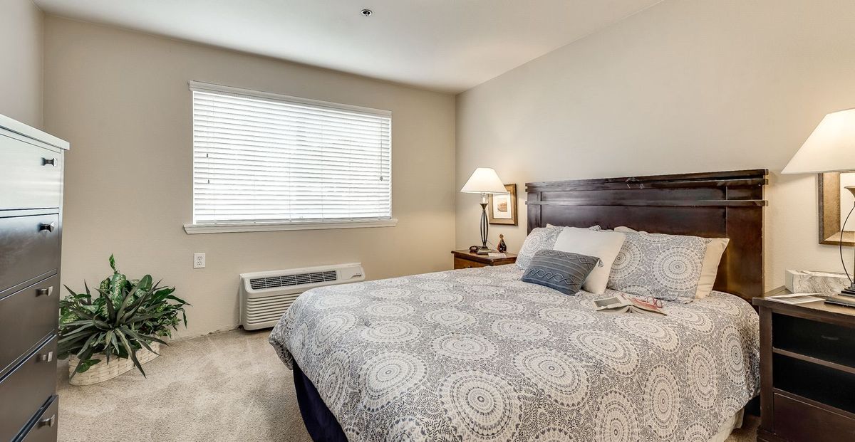 A cozy bedroom with a neatly made bed, bedside lamp, and a window letting in natural light.