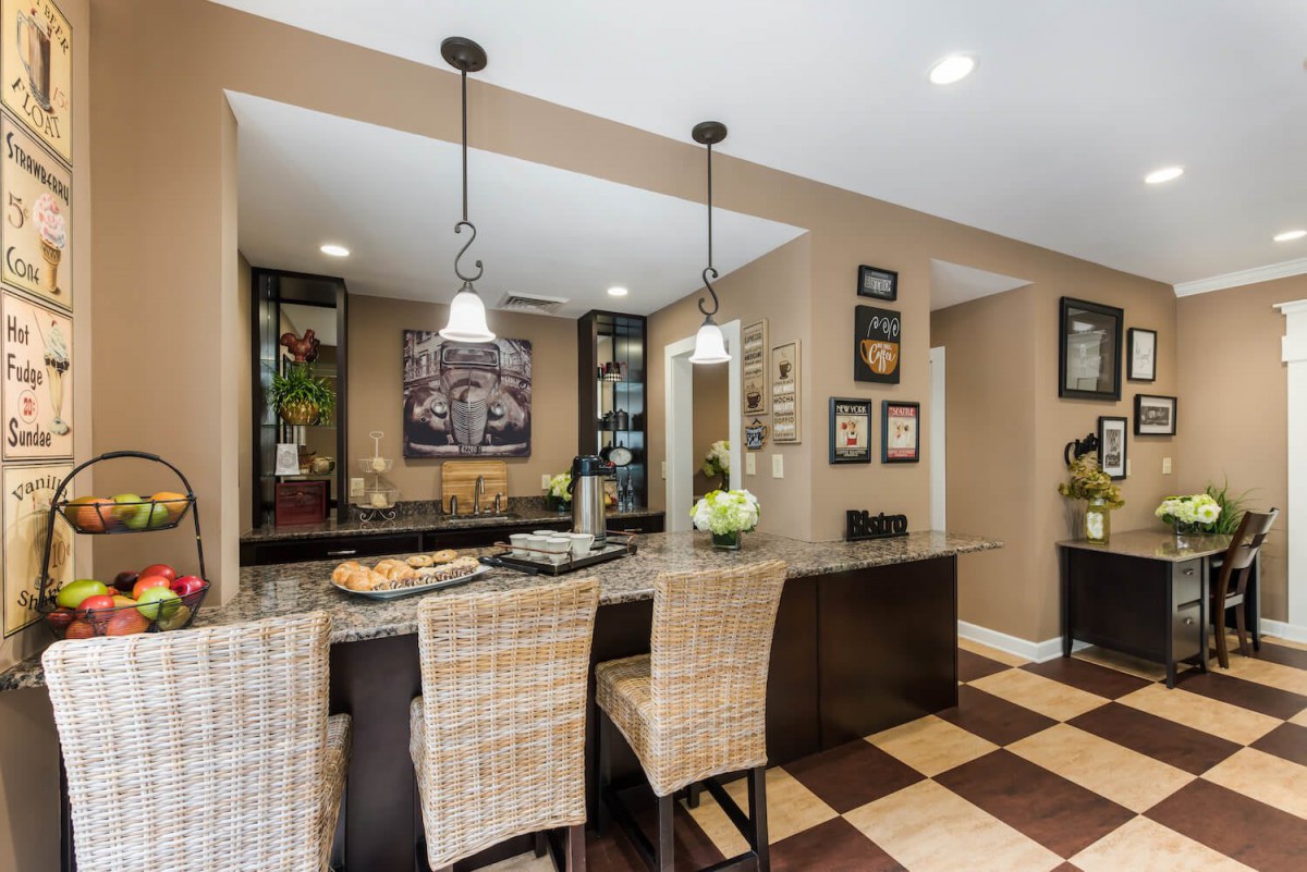 A modern kitchen and dining area featuring pendant lights over a granite countertop with wicker bar stools, enhanced by decorative wall art and a checkered floor.