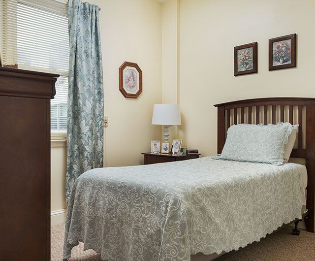 A cozy bedroom featuring a single bed with vintage-style linens, complemented by curtains, a bedside lamp, and framed pictures on the wall.