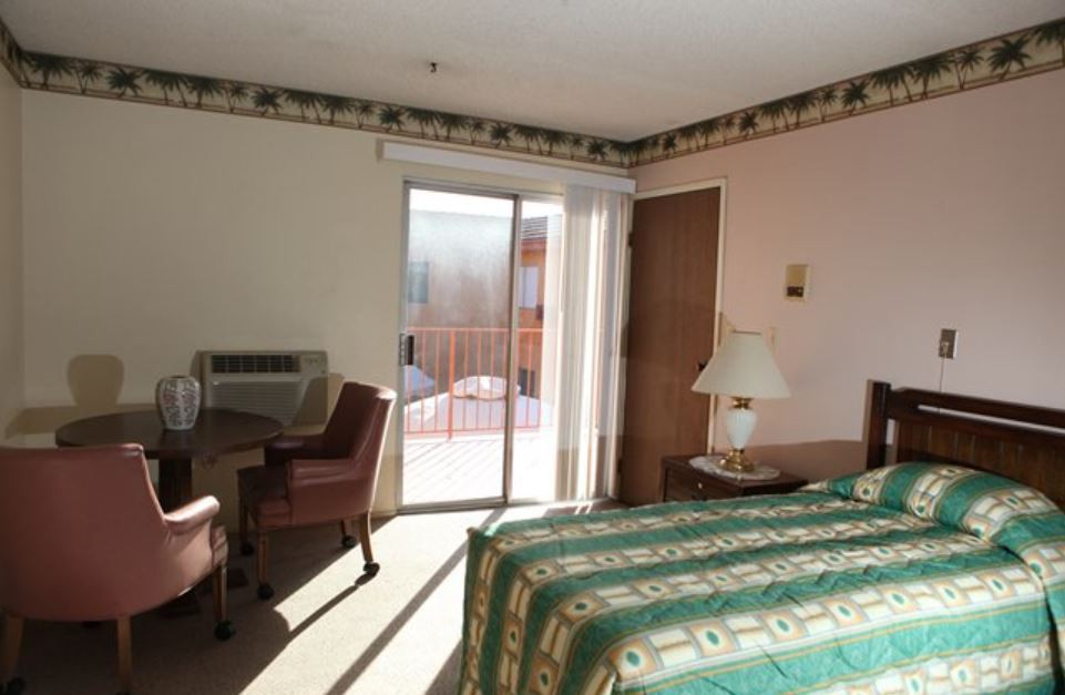 A tidy hotel room with a bed, desk, and sliding glass door letting in natural light.
