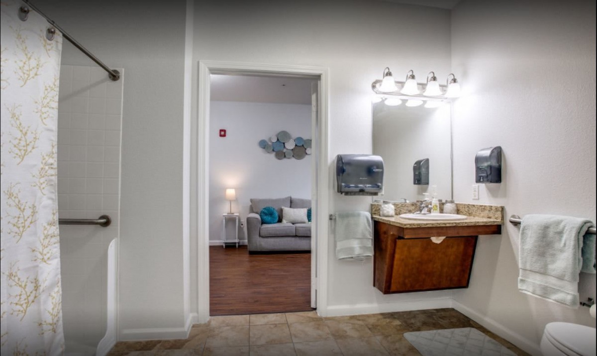 A well-lit bathroom with a view into an adjoining living room featuring a sofa and wall decor.