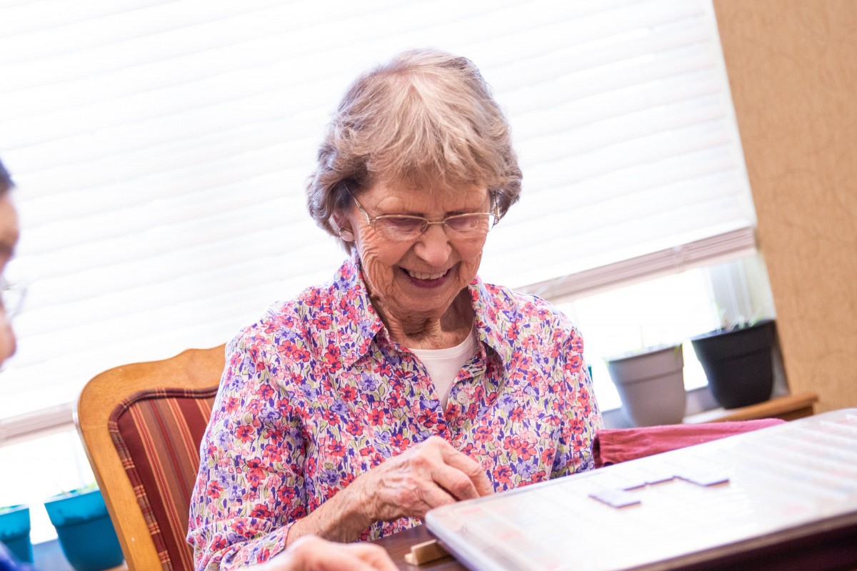 An elderly woman wearing glasses and a floral shirt is smiling indoors.