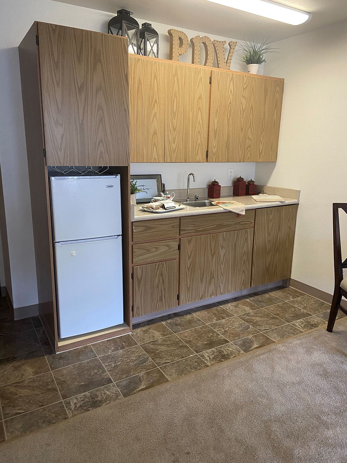 A compact kitchen space featuring wooden cabinets, a small refrigerator, and a sink with tiled flooring.