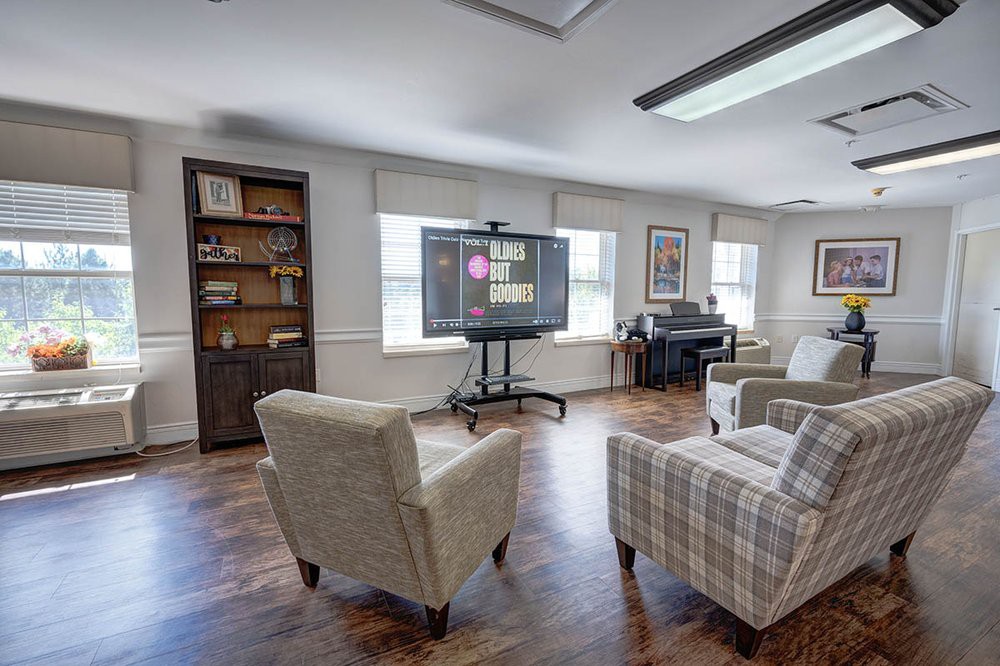A cozy living room with comfortable chairs facing a television, adorned with a bookshelf and large windows letting in natural light.