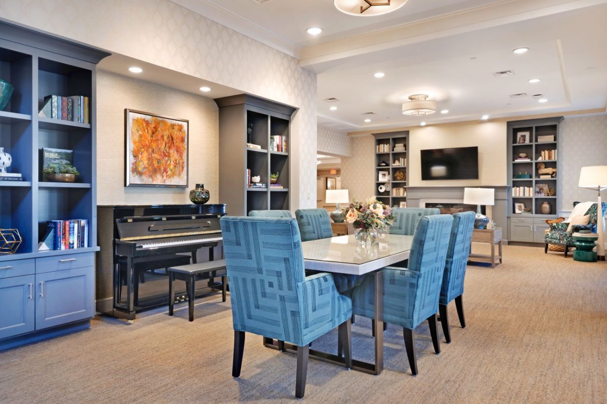A modern living and dining area featuring blue upholstered chairs, a piano, contemporary artwork, and built-in shelves.