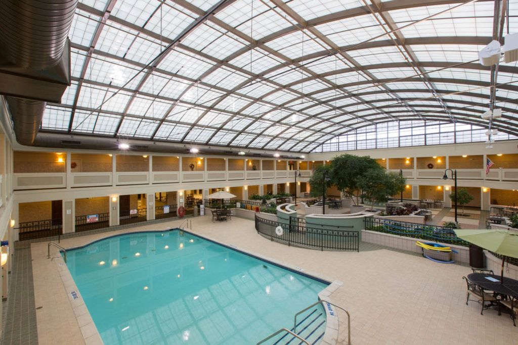 A spacious indoor swimming pool beneath a large glass atrium, surrounded by hotel amenities and seating areas.