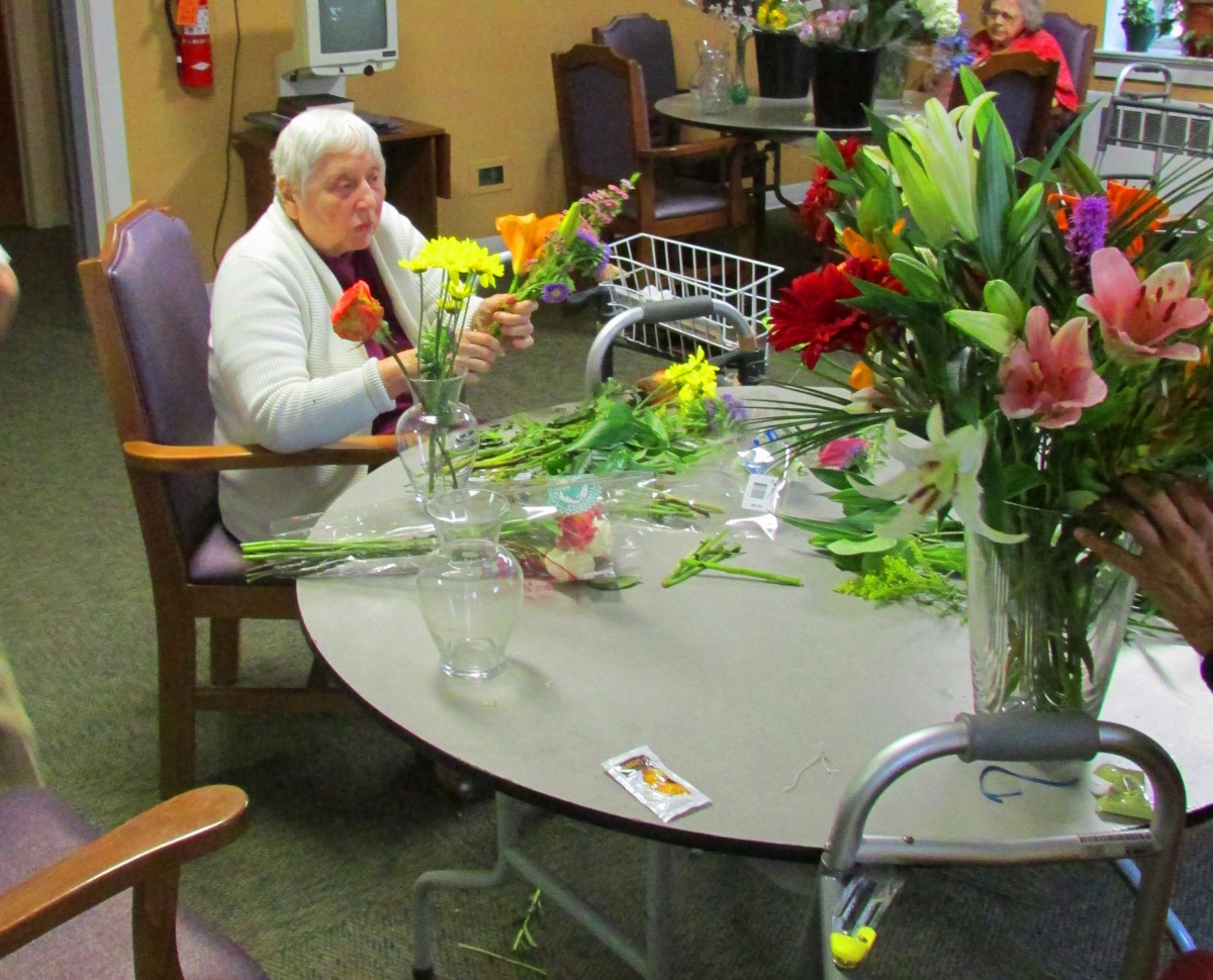 An elderly woman arranges flowers at a table in a retirement home.