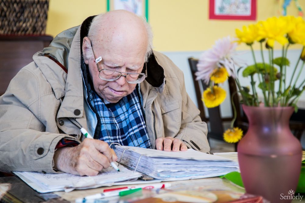 An elderly man deeply focused on writing in a notebook indoors.