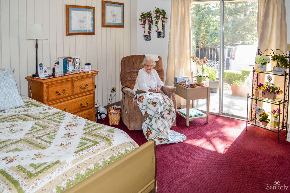 An elderly woman sits comfortably in a cozy bedroom filled with sunlight, surrounded by personal decor and a warm quilt.