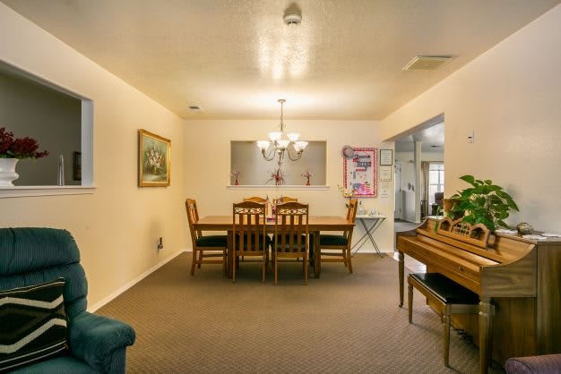 A cozy dining room with a wooden table and chairs, illuminated by a chandelier, featuring artwork on the walls and a piano in the corner.