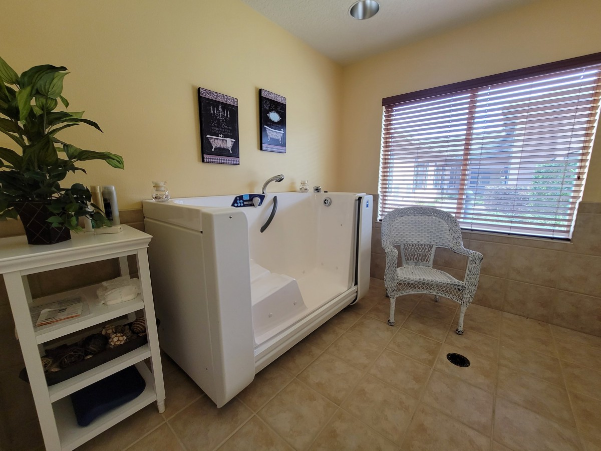 A cozy room featuring a large, accessible bathtub with a nearby chair and shelf, illuminated by natural light from the window.
