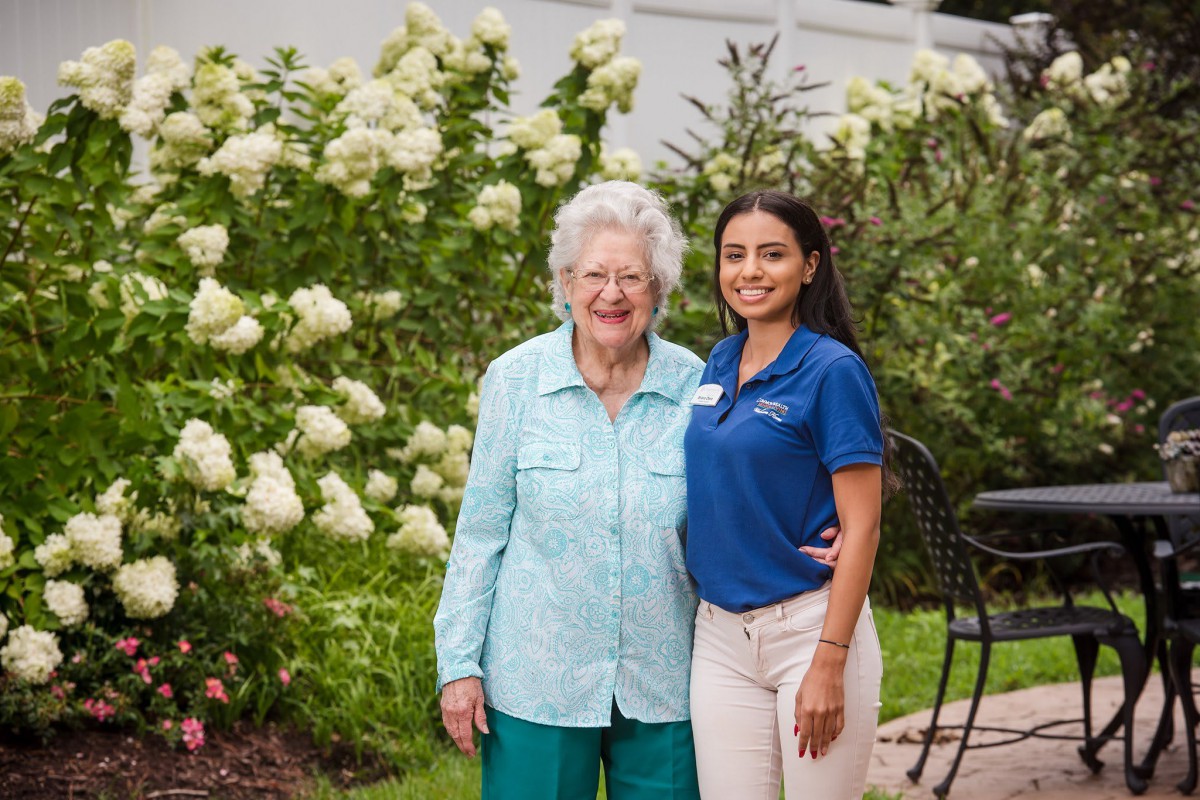 An elderly woman and a younger woman smiling together in a garden full of white flowers.
