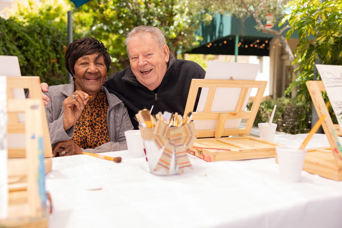 Two seniors enjoying a painting session outdoors, surrounded by art supplies.