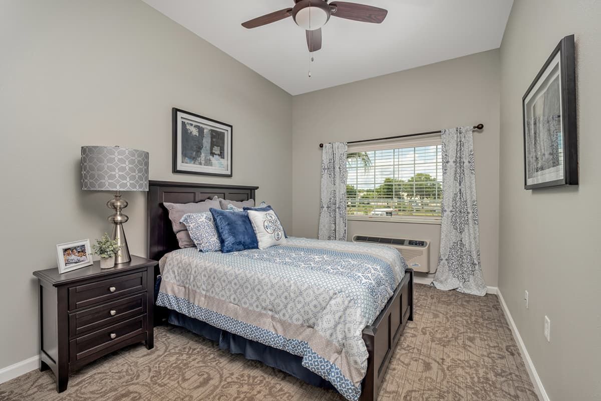 A cozy bedroom with a neatly made double bed, featuring elegant decor and a ceiling fan. A window with curtains allows natural light to fill the room.