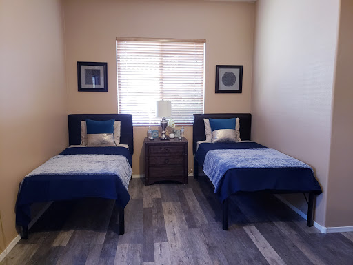 A cozy bedroom featuring twin beds with blue bedding and a nightstand in between, illuminated by natural light from a window.
