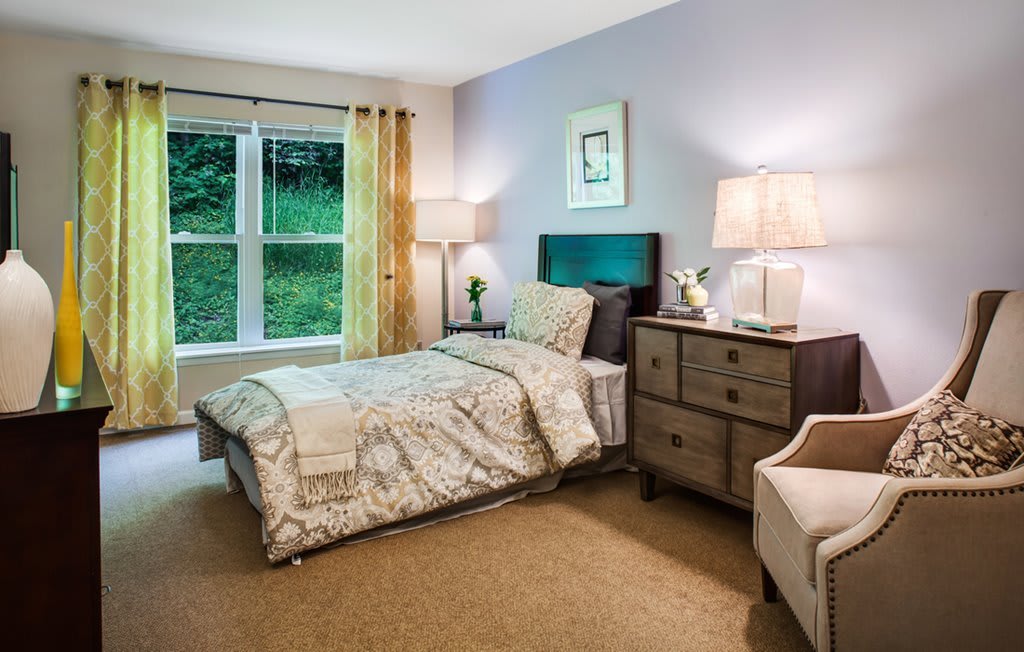 A cozy bedroom with a neatly made bed, side table with lamps, and a large window.