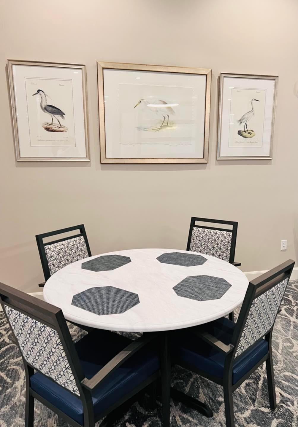 A round dining table with four chairs is set with placemats, and the surrounding wall features framed bird artwork.