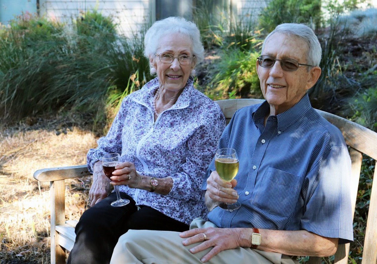 An elderly couple enjoying wine while sitting outdoors in a garden.