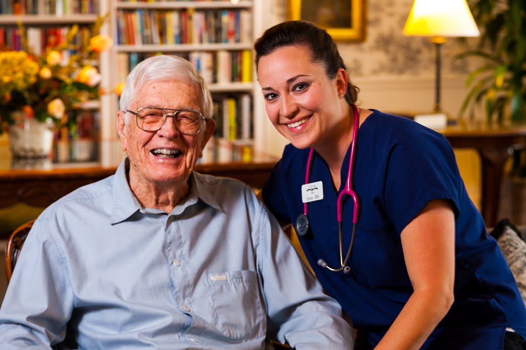 An elderly man sits happily with a nurse in a cozy living room setting, showcasing a warm and caring atmosphere.