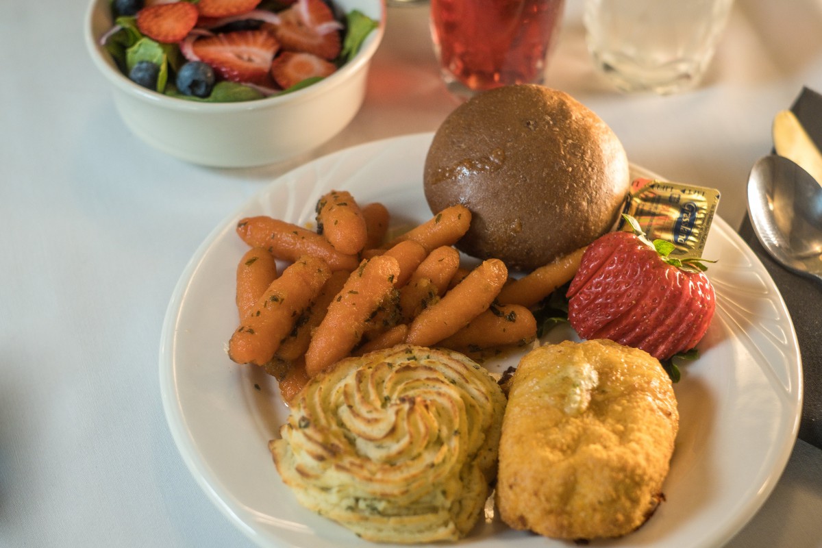 A plate featuring carrots, a bread roll, a swiss roll, a piece of cake, and strawberries with a side salad.