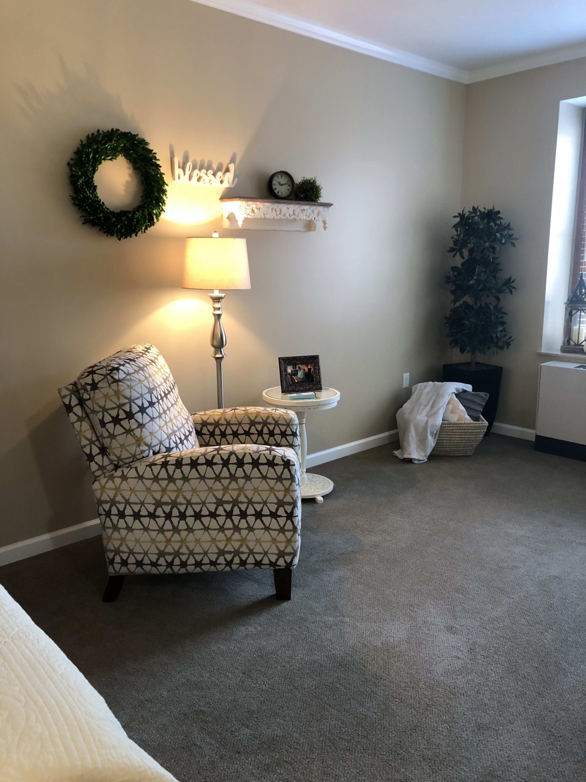 A cozy living room corner with a patterned armchair, floor lamp, decorative wreath, and a small round table.