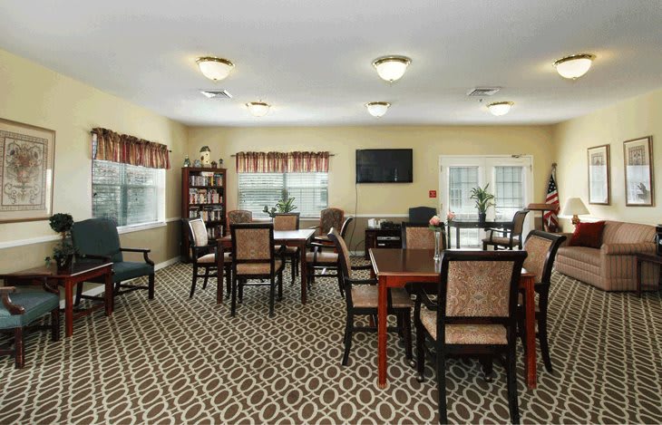 A cozy dining area with tables and chairs, featuring a patterned carpet, windows, and a television.